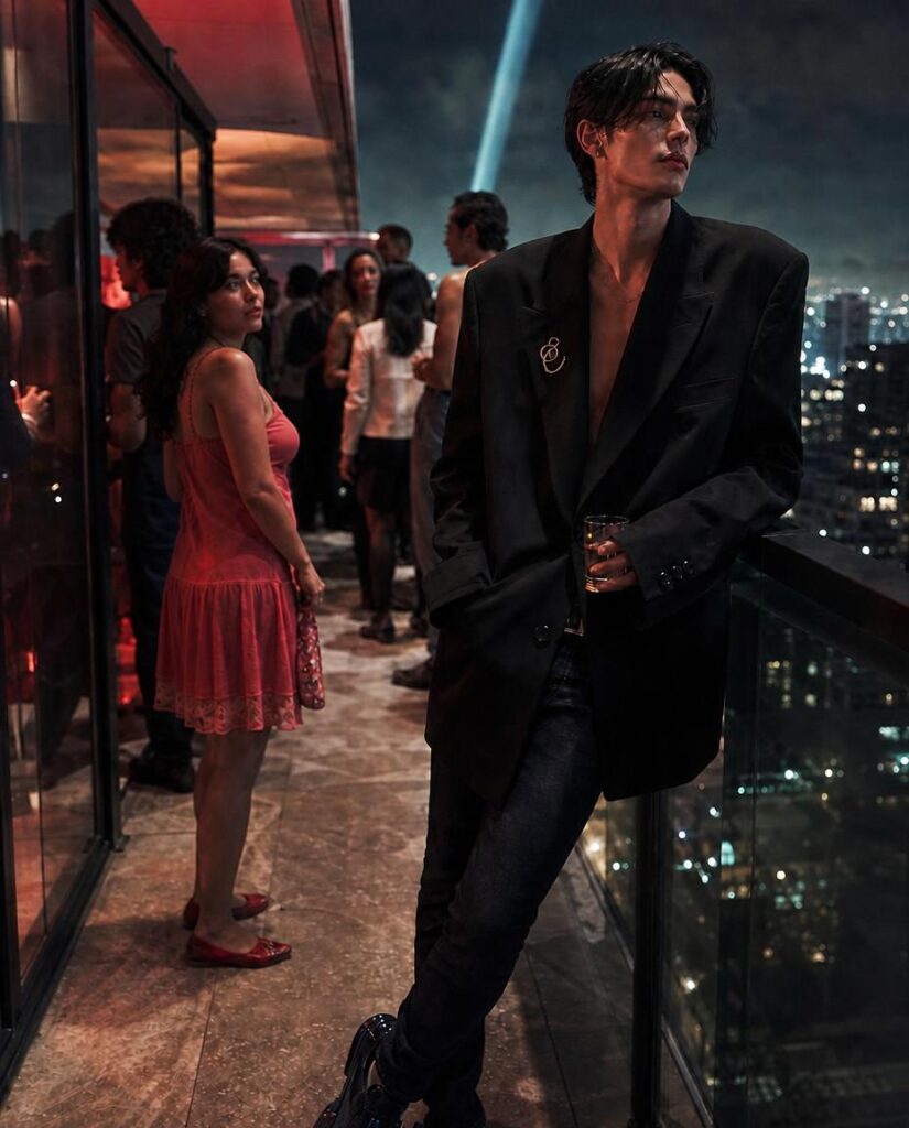 Androgynous young man in an oversized black blazer and fitted denim jeans, leaning against the railing of a luxury apartment terrace during an evening gathering.
